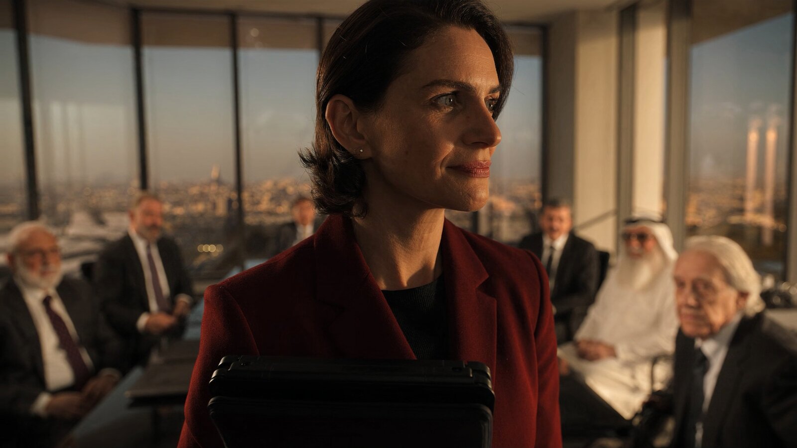 A Lebanese-British woman in a deep burgundy blazer stands thoughtfully in a glass-walled Riyadh boardroom, golden-hour light through floor-to-ceiling windows, Saudi colleagues seated around the table behind her. Soul Cinema cinematic register, no magazine chrome, no masthead.